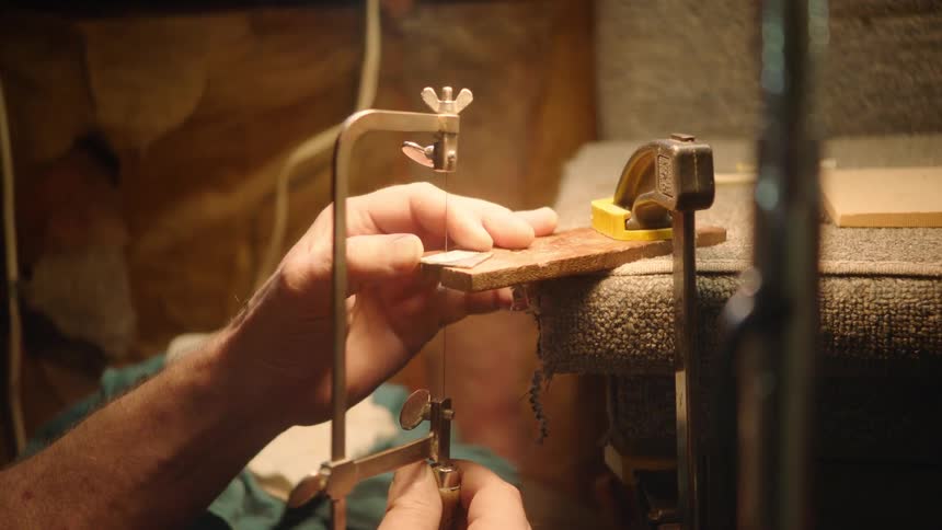 An artisan carefully uses a hand saw to cut a detailed shape from a small piece of material in a warm-lit workshop