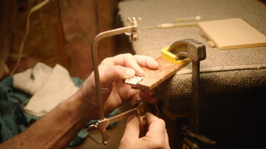 An artisan carefully saws a detailed pattern into a small piece of metal clamped to a workbench in a warmly lit workshop