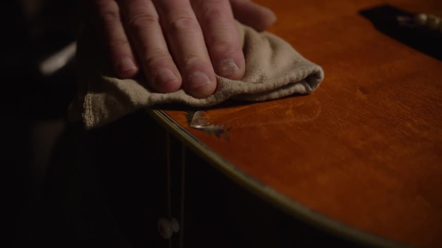A man's hand carefully wipes down the body of a wooden acoustic guitar with a cloth in slow motion