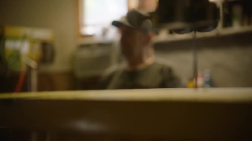 A woodworker in a cap stands thoughtfully in his workshop as the camera rack focuses to a band saw in the foreground