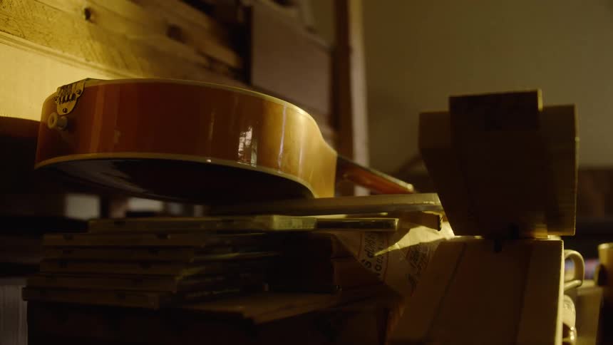 A vintage mandolin rests on a cluttered shelf in a warmly lit, dusty room
