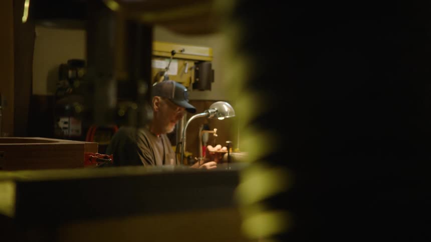A rack focus shot reveals a skilled artisan diligently working on a small craft project in his dimly lit workshop