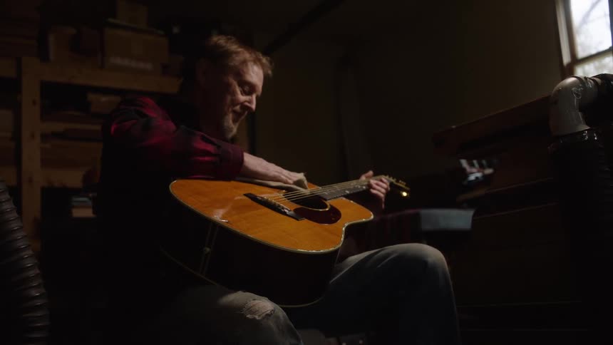 A focused man in a plaid shirt carefully polishes his wooden acoustic guitar in a dimly lit workshop