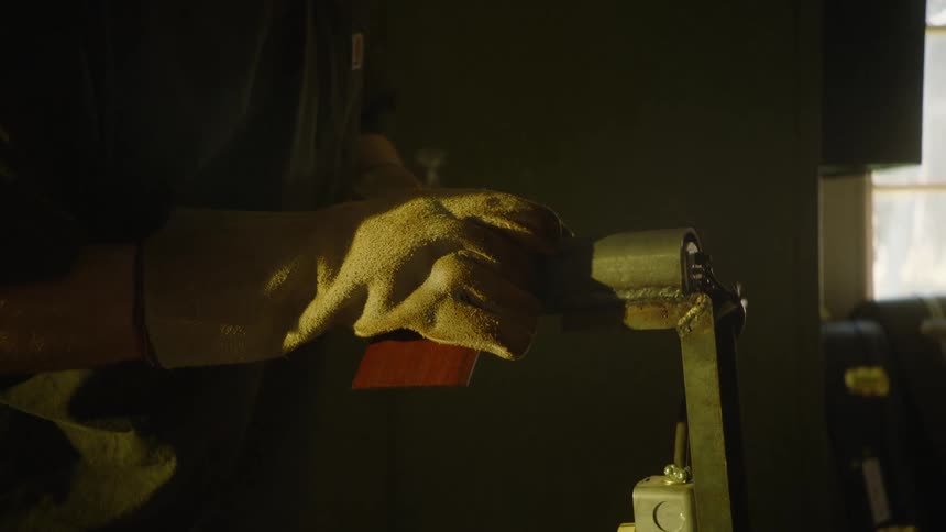 A luthier wearing a protective glove carefully shapes a wooden guitar neck on a belt sander in a dimly lit workshop