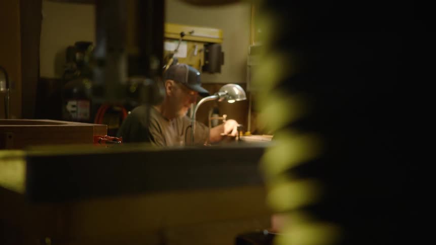 A rack focus from a bandsaw reveals an artisan working diligently at his workbench in a dimly lit workshop