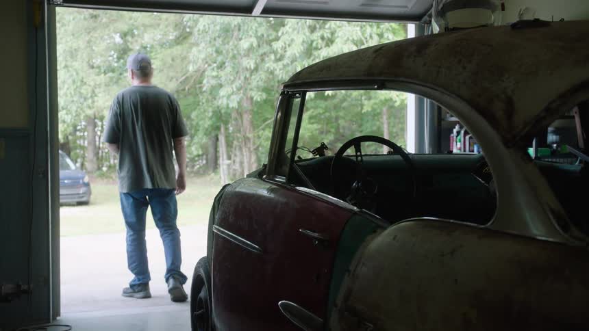 A man slowly walks out of his garage, the silhouette of a classic car restoration project visible inside