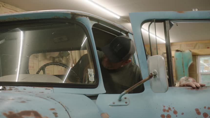An older man opens the door and gets behind the wheel of a classic rusty blue pickup truck in his garage