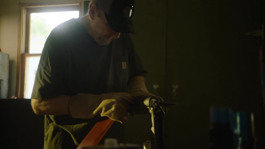 A craftsman carefully sands the edge of a long leather strap in his dimly lit workshop