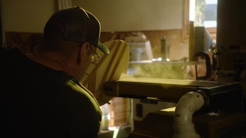 A woodworker carefully sands a custom board on a belt sander in slow motion, with sawdust particles filling the air in his workshop