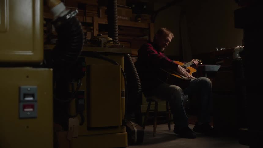 A luthier sits in his dimly lit workshop carefully cleaning and polishing a handcrafted acoustic guitar
