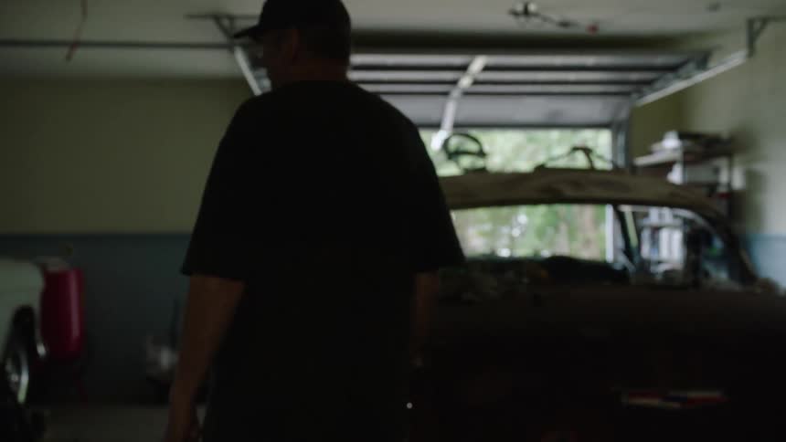 A man in a t-shirt and cap walks slowly into a garage with a vintage car project inside