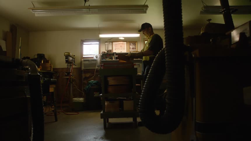 A woodworker enters his shadowy, well-equipped workshop and begins preparing a piece of wood at his workbench