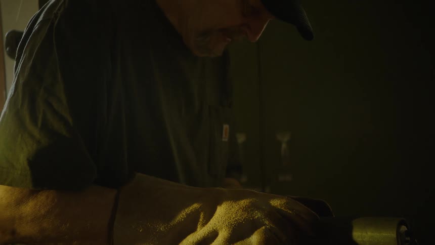A focused craftsman sands a piece of wood with a belt sander in a moody, dark workshop
