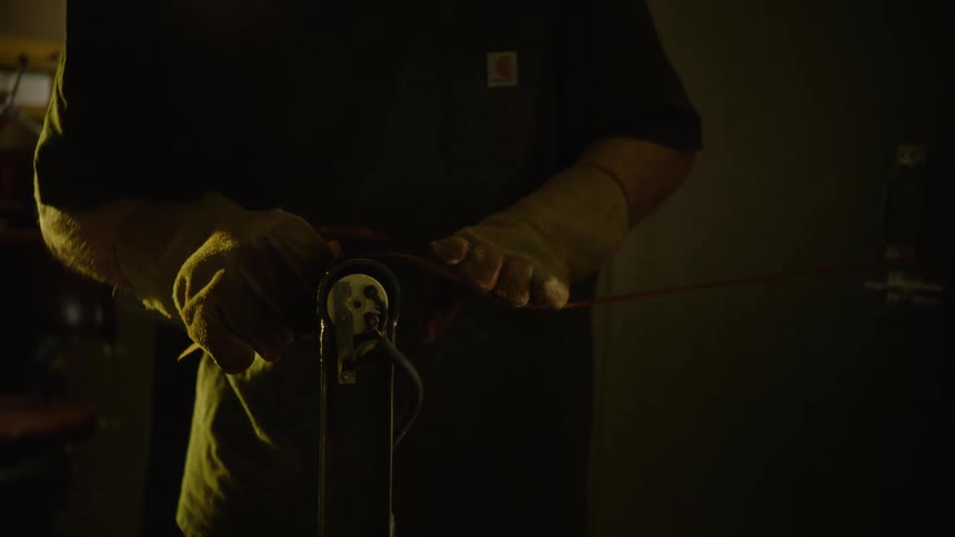 A craftsman wearing work gloves carefully sands the edges of a leather belt on a belt sander in a dimly lit workshop