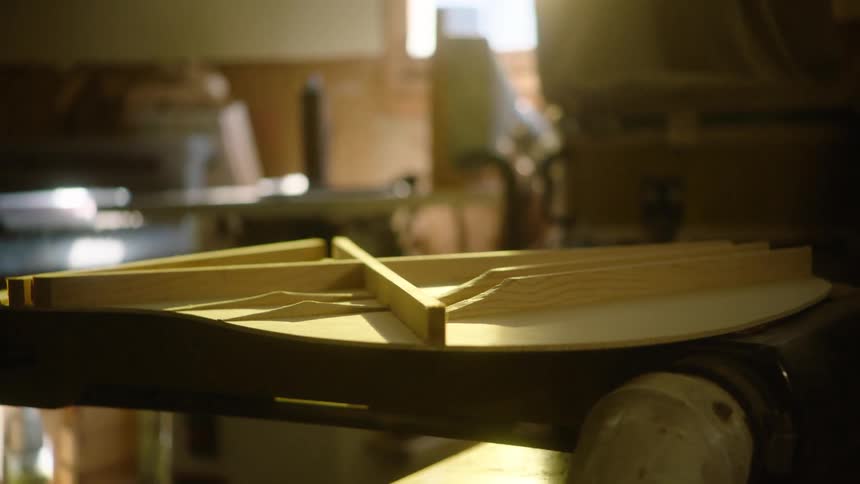 The intricate wooden bracing for an acoustic guitar rests on a workbench in a dusty, sunlit luthier's workshop
