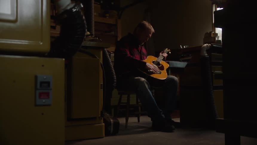 A luthier sits in his dimly lit workshop, carefully sanding and then strumming a beautiful handcrafted acoustic guitar