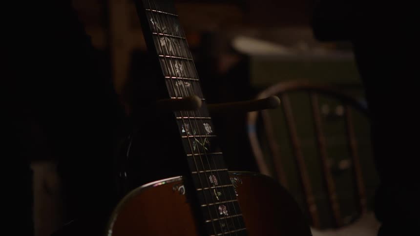 Close up of a musician setting a beautiful acoustic guitar with floral inlays onto a stand in a dark, moody room