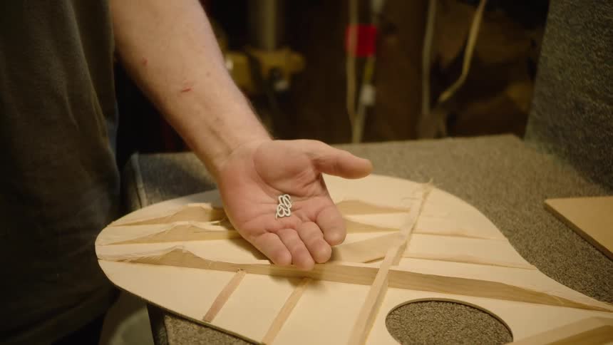 A close-up shot shows a luthier's hand holding small metal hooks above the braced wooden top of a guitar in a workshop