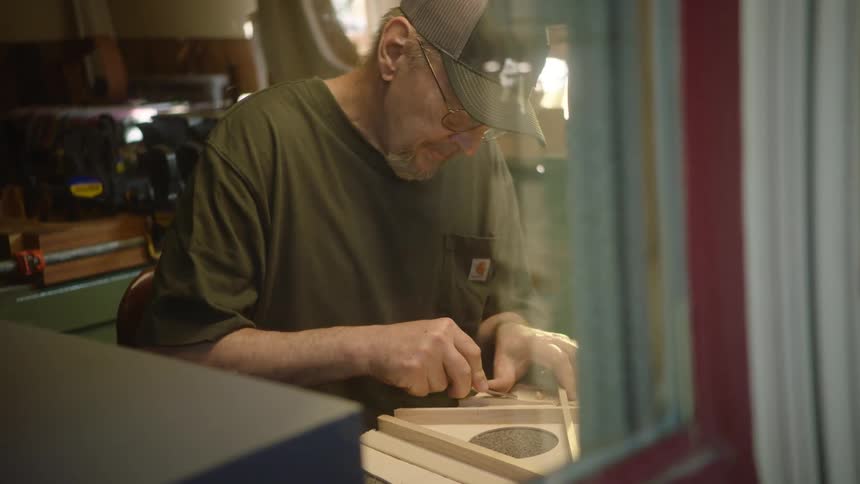 Viewed through a window, a dedicated craftsman meticulously carves and shapes a wooden frame in his workshop
