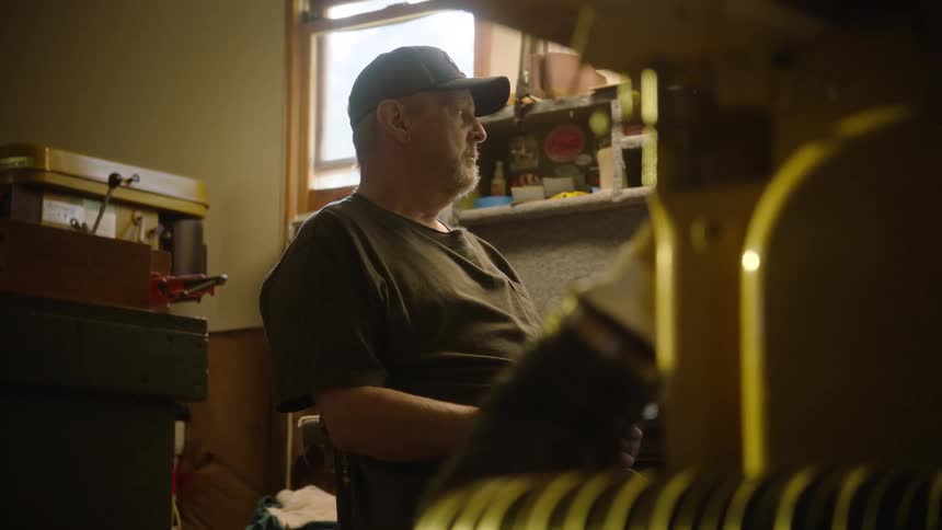 A pensive middle-aged man wearing a baseball cap sits alone in his dimly lit workshop, surrounded by tools
