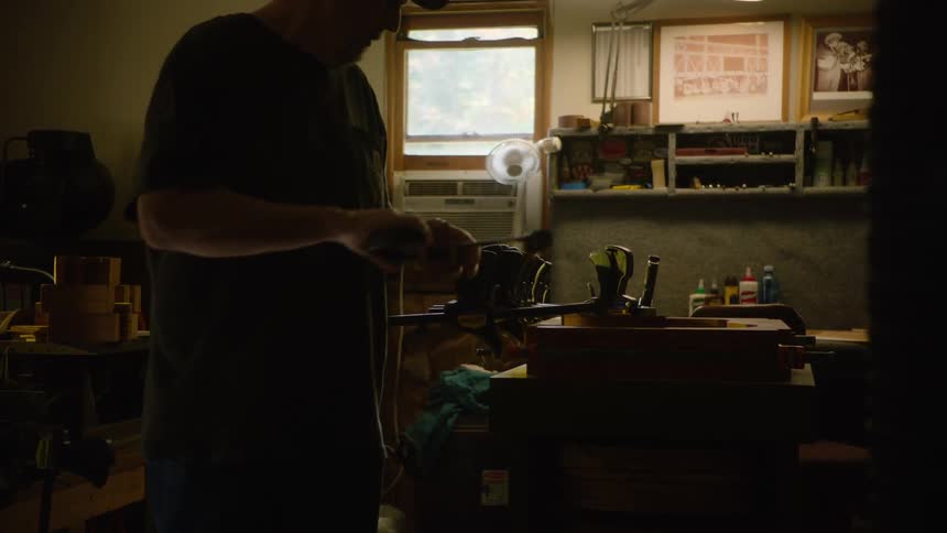 A male craftsman in a dimly lit workshop carefully adjusts clamps on a piece of wood at his workbench