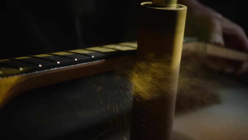 Close-up in slow motion of a luthier using a spindle sander to shape a wooden guitar neck, with fine sawdust flying through the air