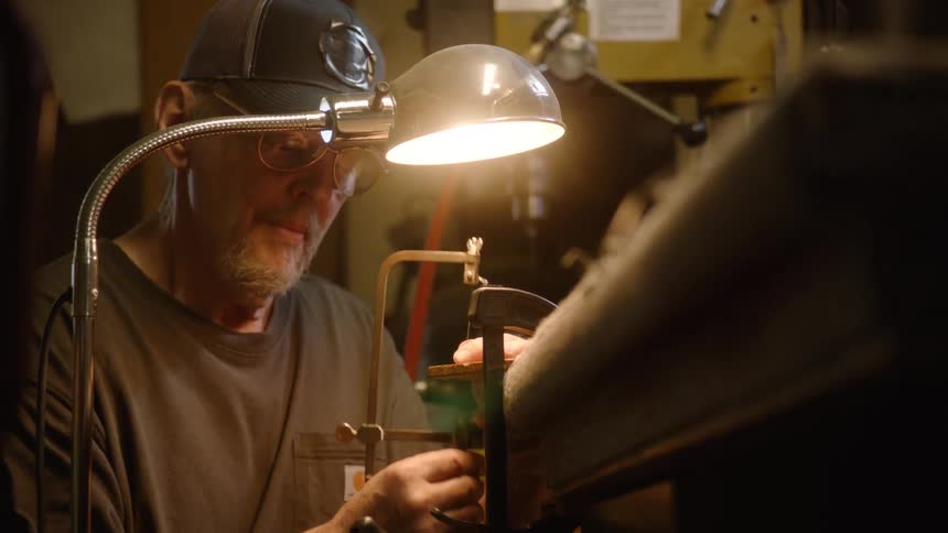 An older craftsman meticulously saws a small piece of material at his workbench, illuminated by a single focused lamp in his workshop