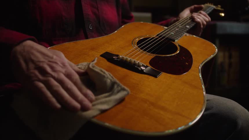 A craftsman in a dimly lit workshop meticulously polishes the body of a wooden acoustic guitar in slow motion