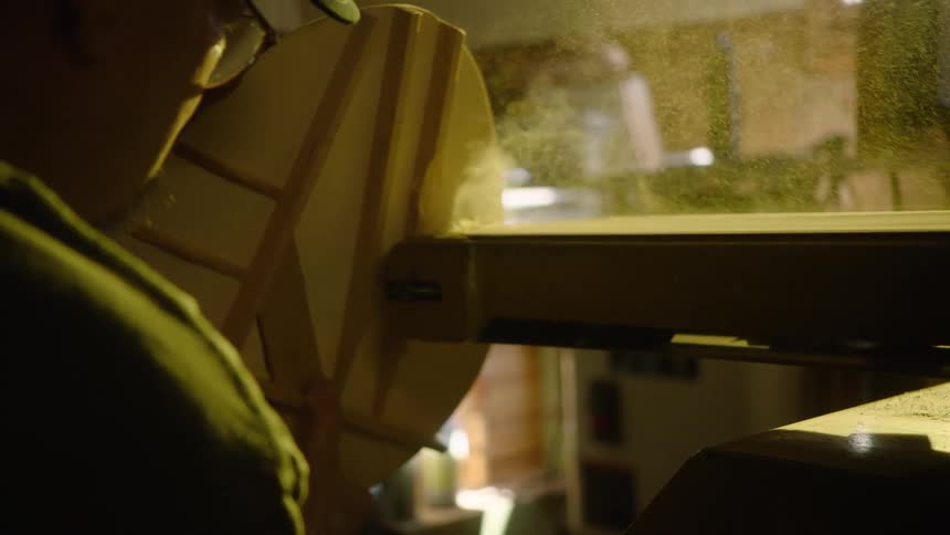 A woodworker carefully shapes a piece of wood on a belt sander, sending a cloud of fine sawdust into the air in slow motion