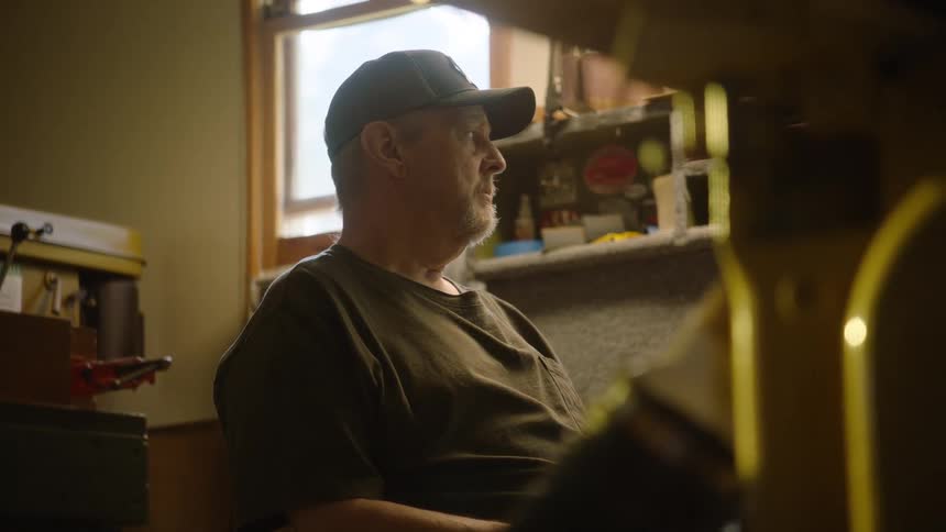 A thoughtful man wearing a baseball cap sits and rests in his home workshop