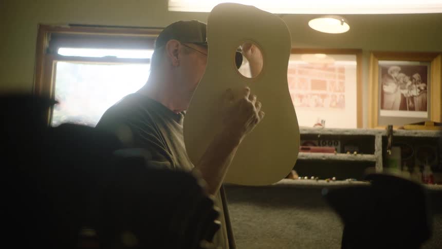 An artisan guitar maker holds up and inspects the wooden top of a guitar in his workshop