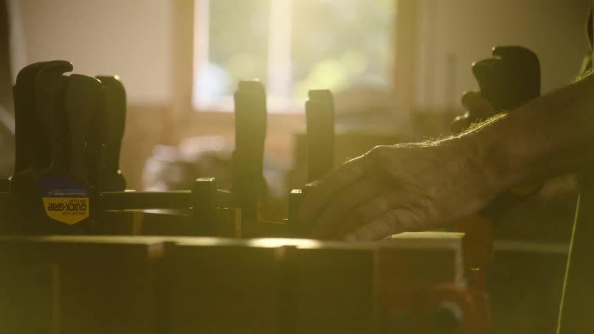 A carpenter carefully tightens clamps on a woodworking project amidst a golden haze in a sun-drenched workshop