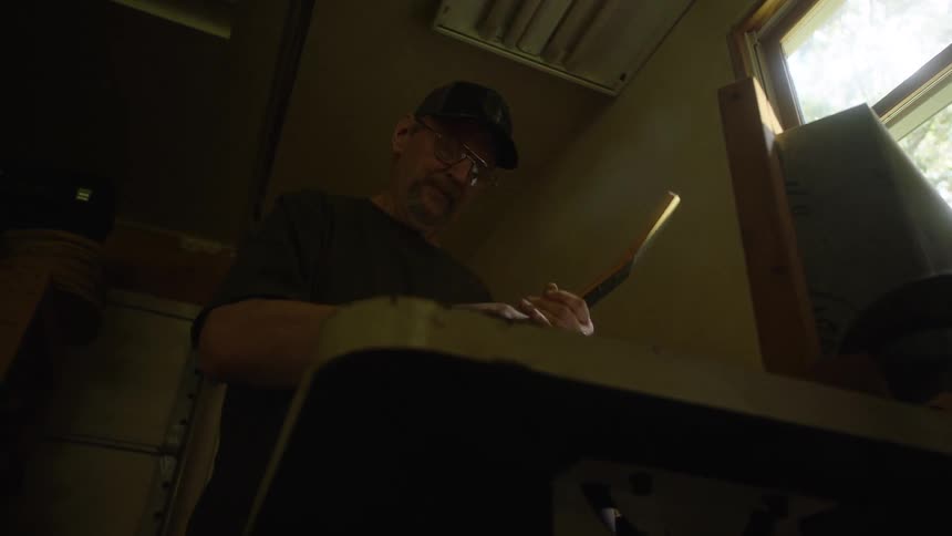 A skilled craftsman in a dimly lit workshop carefully sands the neck of a guitar, with sawdust illuminated by his headlamp