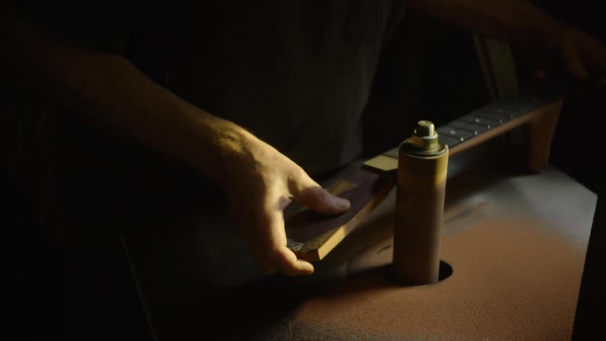 A skilled luthier meticulously shapes and sands the neck of a guitar using a power sander in a workshop