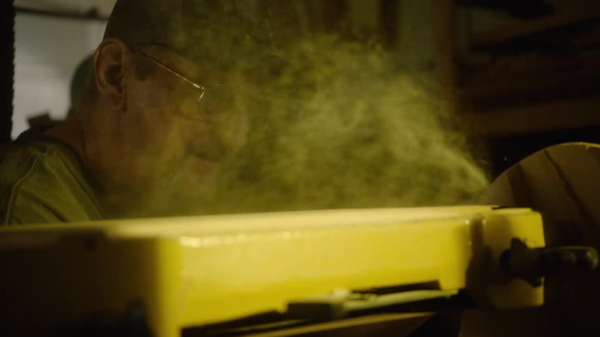A carpenter in a dimly lit workshop uses a power sander on wood, creating a cloud of fine sawdust in dramatic slow motion