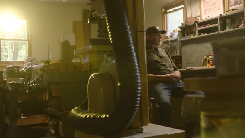 An older man sits thoughtfully in his sun-drenched, cluttered workshop, taking a quiet break from his work