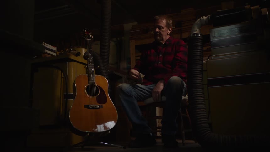 A luthier in his dimly lit workshop proudly inspects his handcrafted acoustic guitar before placing it on a stand and walking away