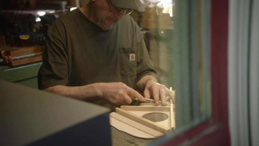 A luthier seen through a window meticulously carves the interior bracing of a handmade acoustic guitar in his workshop