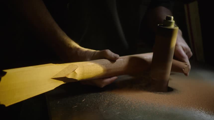 A craftsman meticulously shapes a wooden guitar neck on a spindle sander, with sawdust flying in dramatic slow motion