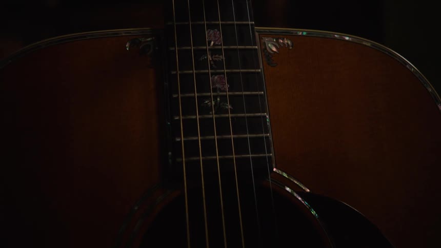 A close-up shot slowly pans down the neck and body of a beautifully crafted acoustic guitar, highlighting the intricate floral inlay