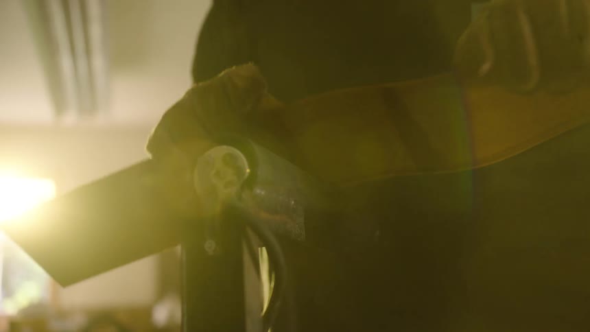 A craftsman wearing gloves carefully feeds a leather strap into a sanding machine in a warm, sunlit workshop