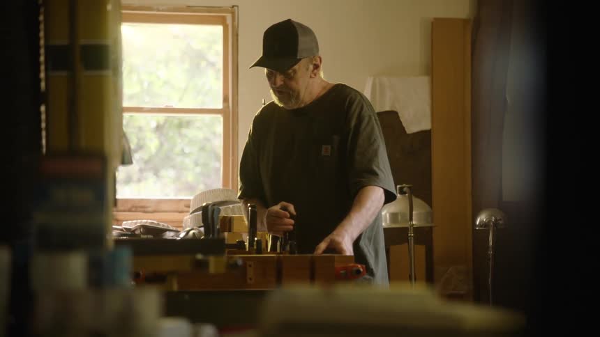 A skilled craftsman meticulously works at his bench in a sunlit workshop, framed by tools and equipment