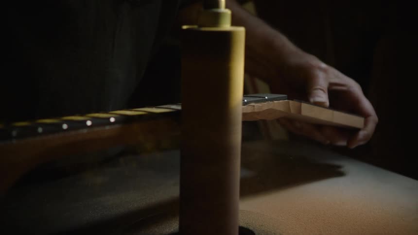 A skilled luthier meticulously shapes the neck of a guitar using a power sander in a dimly lit workshop, captured in slow motion