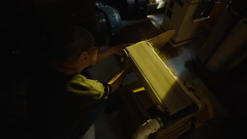 Overhead slow motion shot of a craftsman in a dimly lit workshop using a large belt sander to shape the body of a wooden acoustic guitar