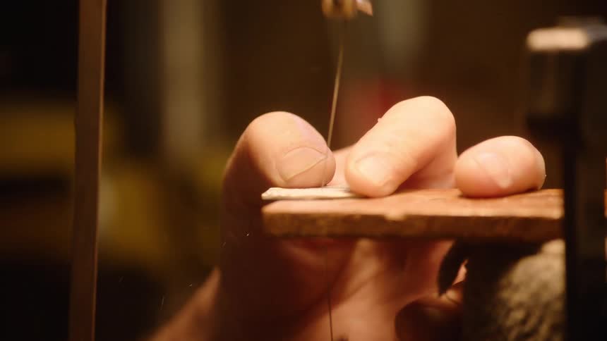 A close-up of a jeweler's hands carefully using a piercing saw to cut a thin piece of metal in a workshop