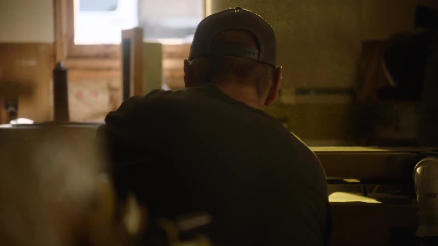 Slow motion shot from behind a man in a workshop as he sands a piece of wood, creating a cloud of illuminated sawdust