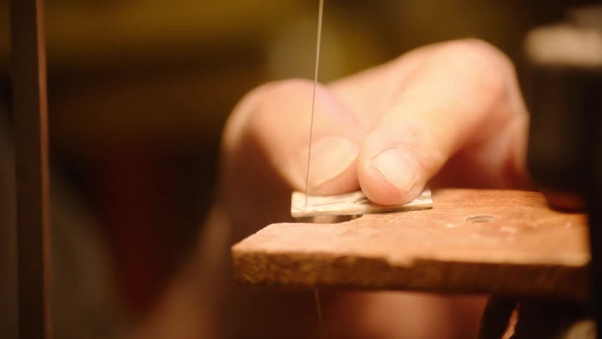 A close-up slow-motion shot captures a skilled jeweler precisely cutting a small piece of metal with a fine saw in their workshop