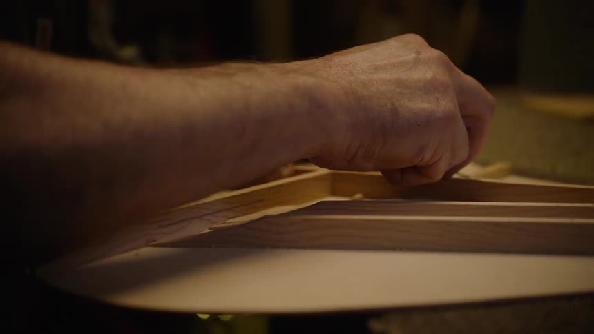 An artisan luthier carefully scrapes away excess glue from the intricate wooden soundboard bracing of a custom acoustic guitar in a workshop
