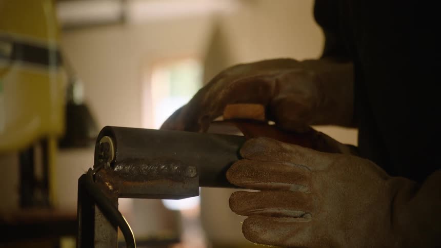 A craftsman in work gloves carefully sands a welded metal pipe on a belt sander in a sunlit workshop