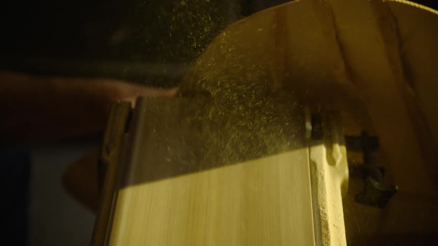 A luthier sands the body of a guitar on a belt sander in slow motion, creating a cloud of fine sawdust